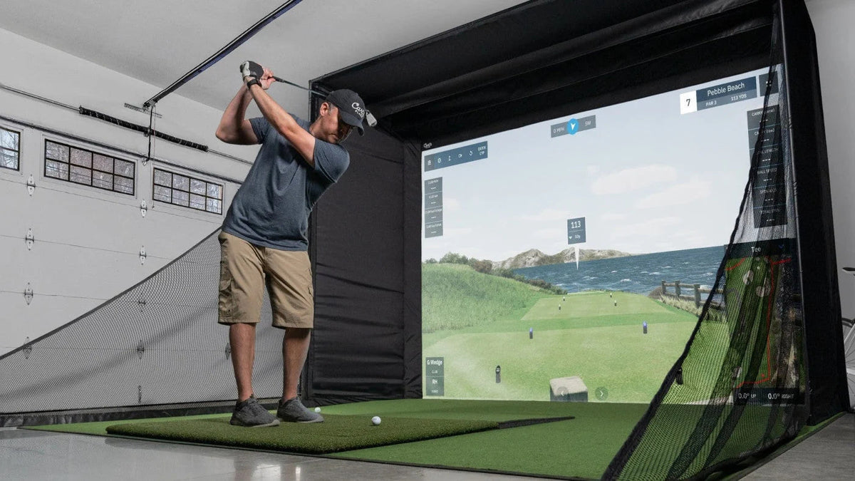 Golfer practicing in a garage golf simulator with an impact screen, hitting mat, and side barrier nets.